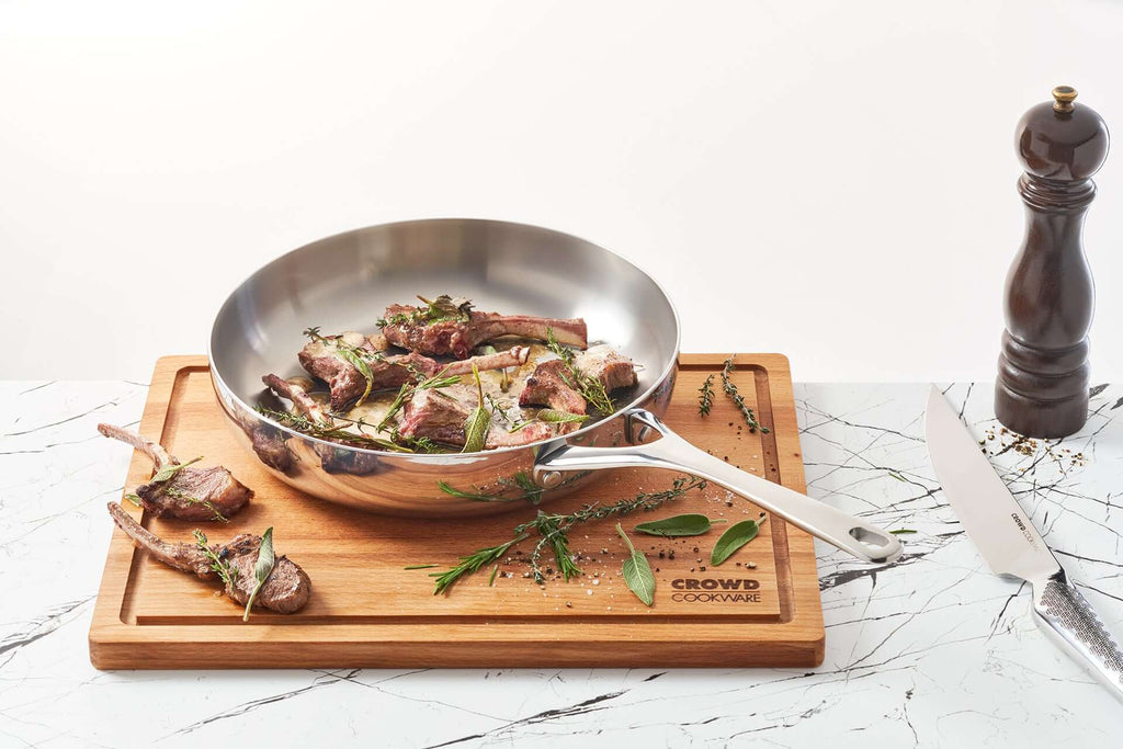 Beech wood cutting board 40x30 cm with integrated juice groove, featuring a 28 cm titanium frying pan with cooked lamb chops, herbs, and Crowd Cookware logo in the corner. A pepper grinder and chef’s knife are placed next to the cutting board o