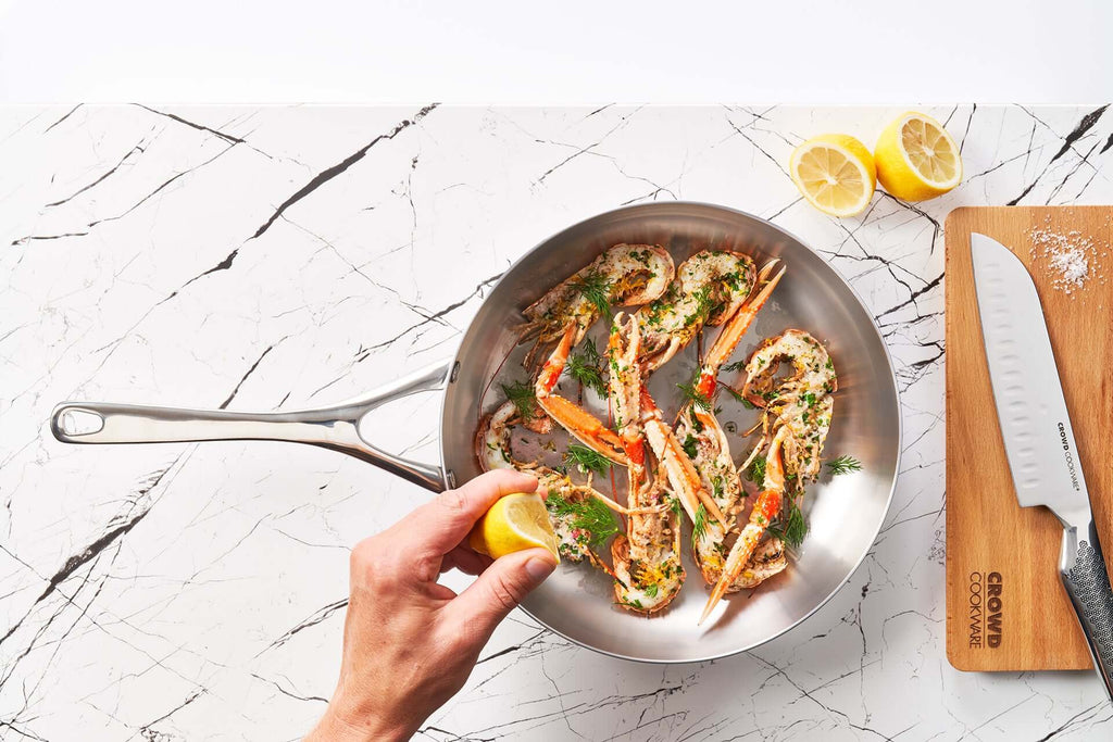 Crowd Cookware 28 cm titanium frying pan with cooked langoustines garnished with herbs, being squeezed with a lemon by hand. A wooden cutting board with a Crowd Cookware chef’s knife and lemon halves are placed on a marble countertop